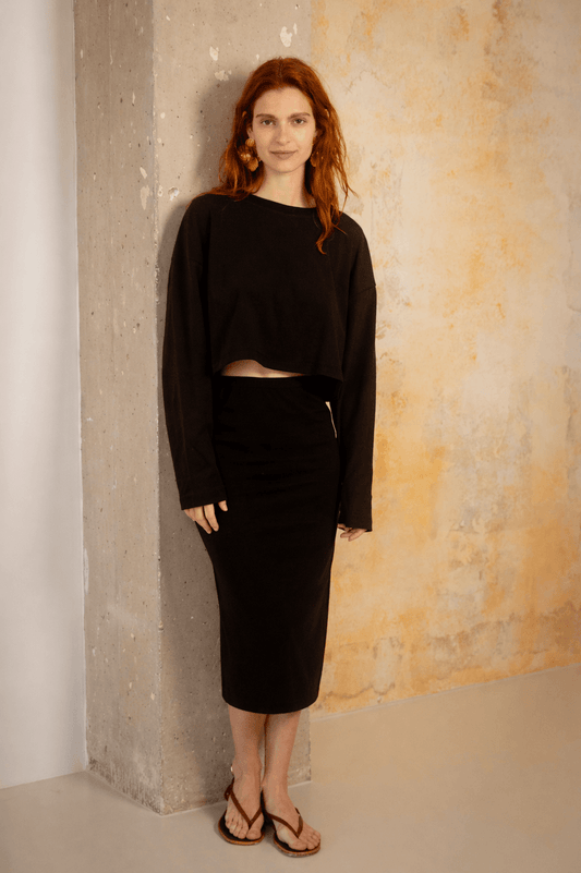 Model wearing Organic Cotton Long Sleeve Top in Black with a black skirt against a textured wall.