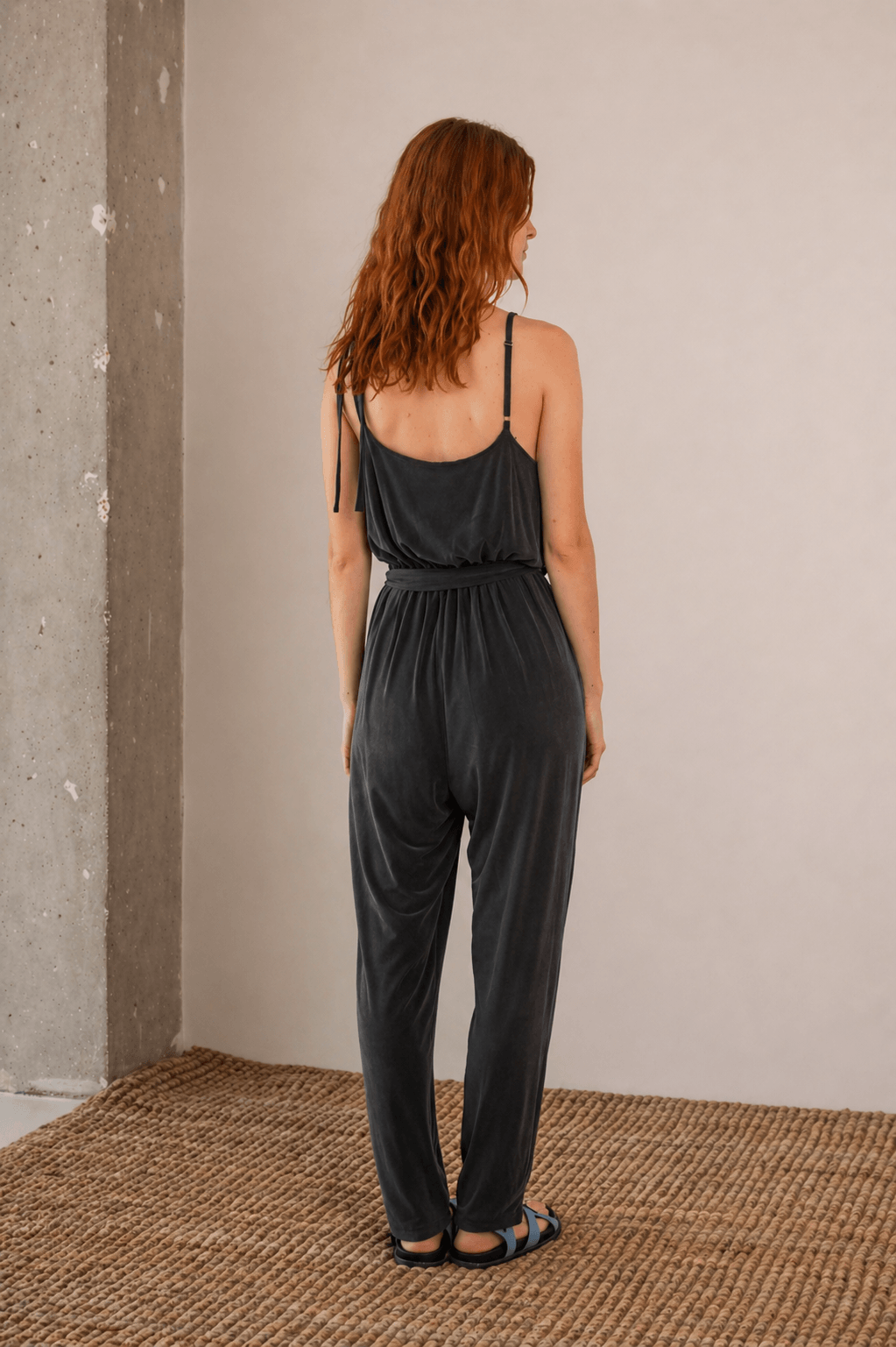 Back view of a model wearing a Cupro Slip Jumpsuit with Belt in Slate Black, showcasing its elegant design.