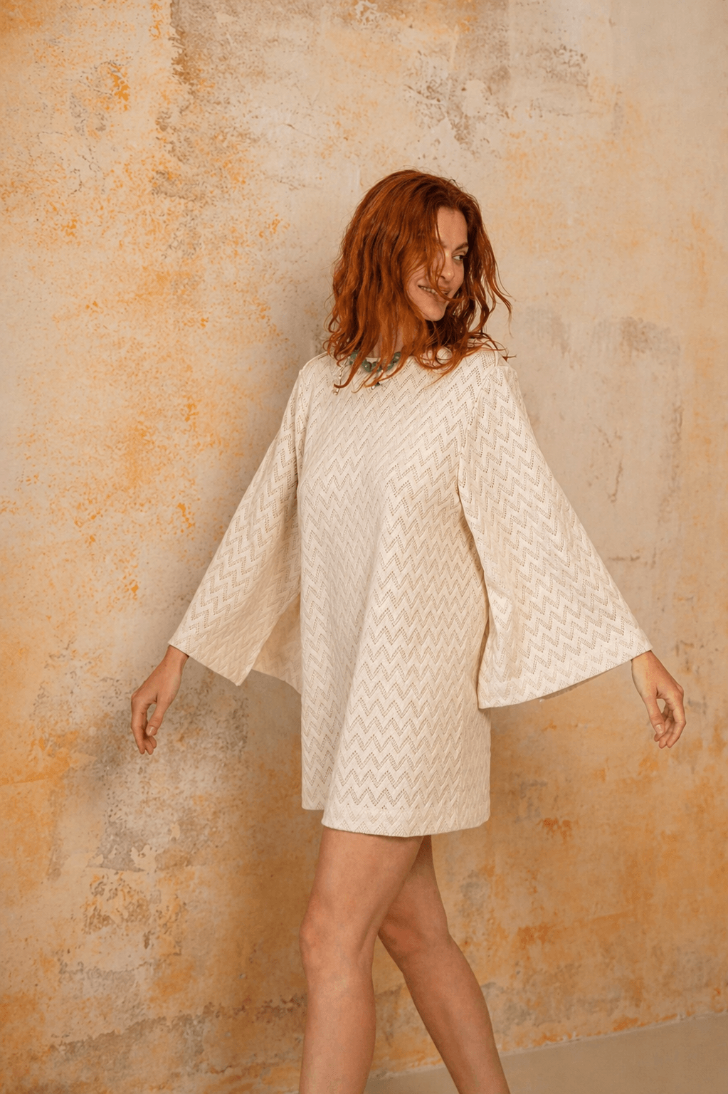 Model wearing an Organic Cotton Crochet Mini Dress with flared sleeves against a textured background.