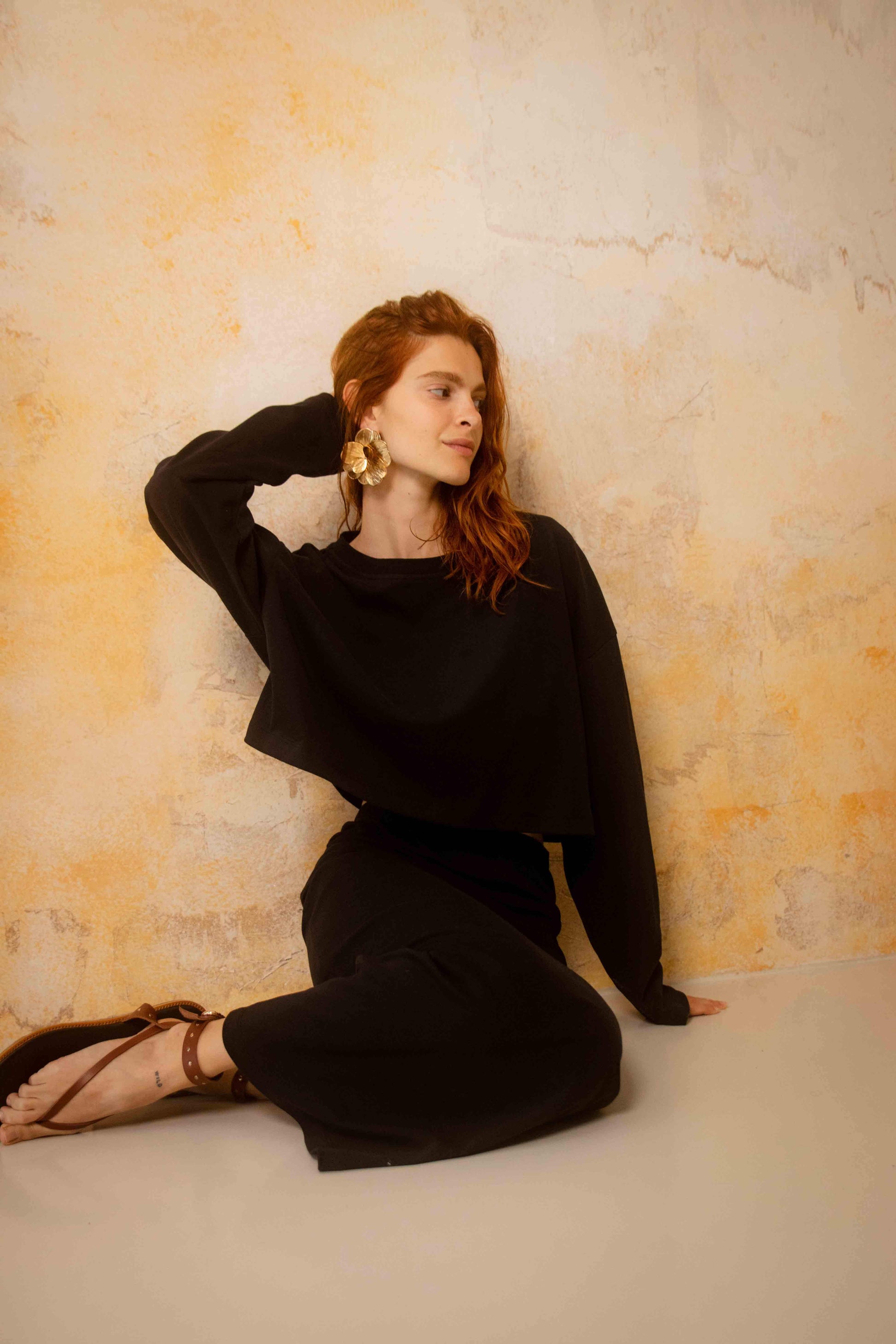 Model wearing Organic Cotton Midi Skirt in Black with a stylish black crop top, seated against a textured wall.