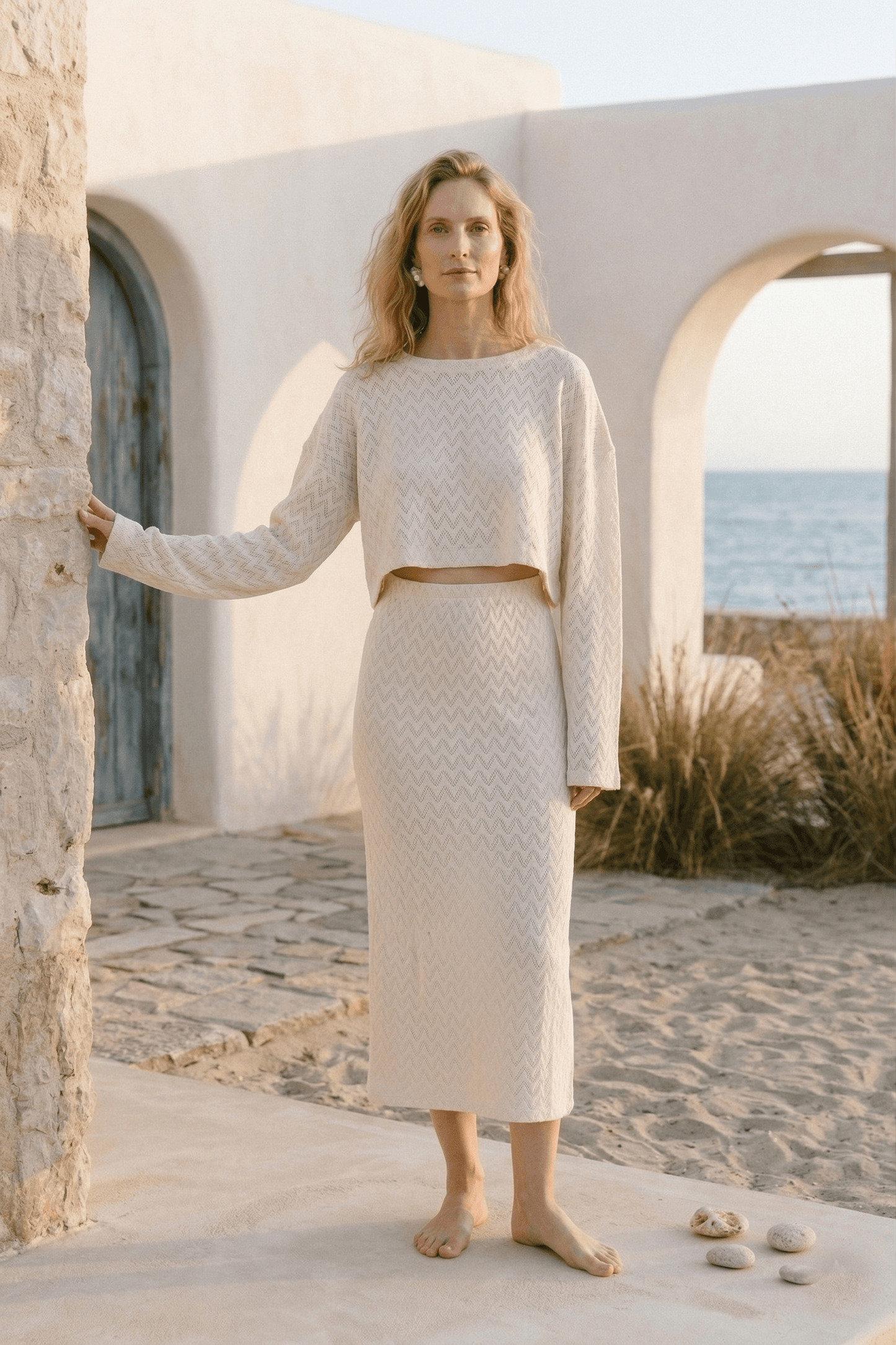Model wearing a Crochet Organic Cotton Midi Skirt and matching top in ecru, with a beach background.