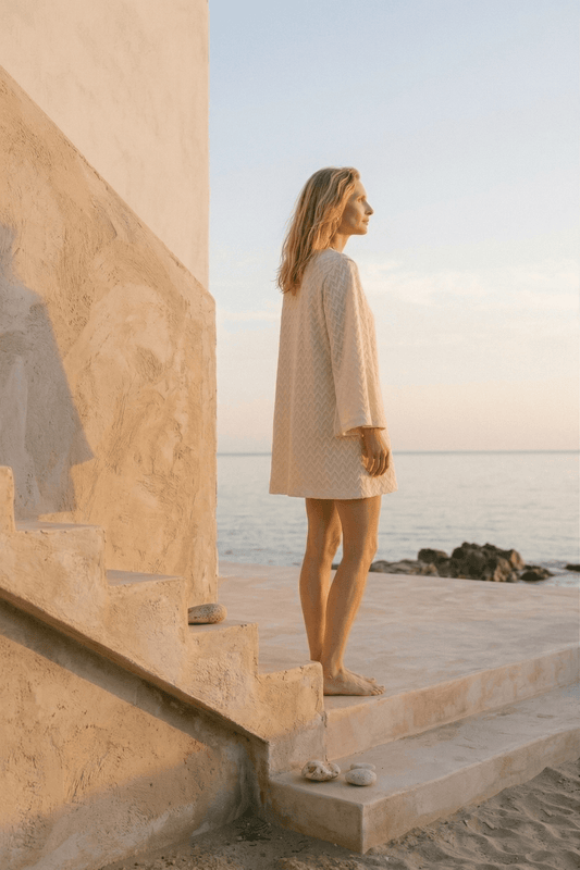 Model wearing Organic Cotton Crochet Mini Dress while standing by the sea at sunset, showcasing its light and elegant design.