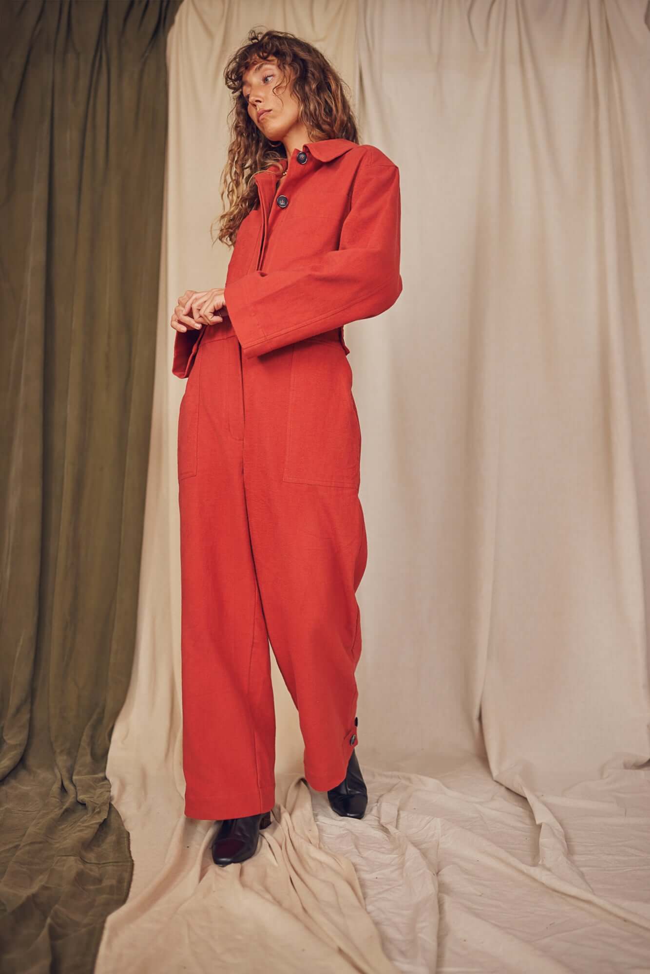 Model showcasing the Rust Organic Cotton Boiler Suit against a backdrop of textured fabrics.